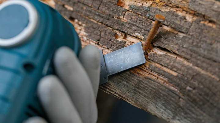 Orang yang mengenakan peralatan keselamatan memotong kayu dengan alat listrik di dekat paku berkarat.