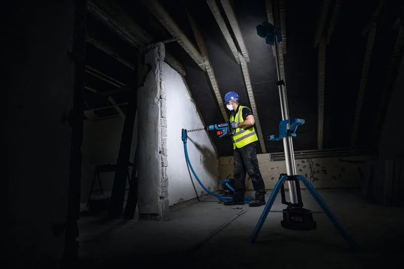 Seseorang yang mengenakan peralatan keselamatan mengebor dinding, diterangi oleh lampu lokasi kerja tanpa kabel.