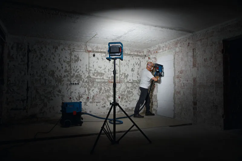 Seseorang yang mengenakan peralatan keselamatan mengoperasikan alat pengeboran dinding di bawah lampu kerja.