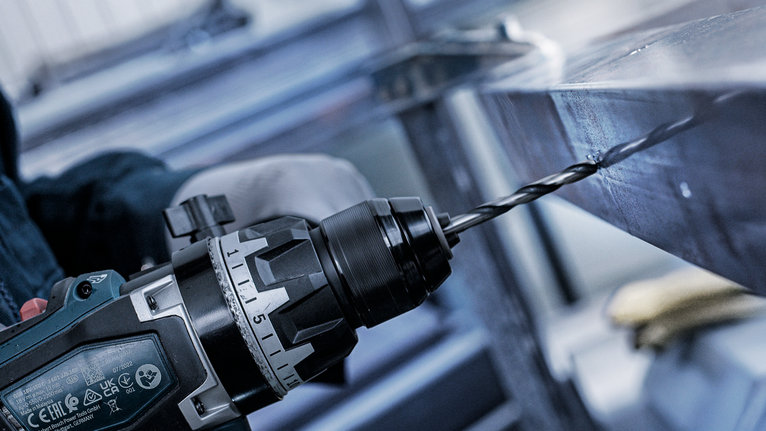 Seseorang yang mengenakan perlengkapan keselamatan mengebor lubang pada balok logam.