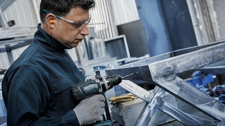 Seseorang yang mengenakan perlengkapan keselamatan mengebor rangka logam.