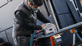 Orang yang mengenakan peralatan keselamatan mengoperasikan gergaji bundar untuk memotong selembar bahan.