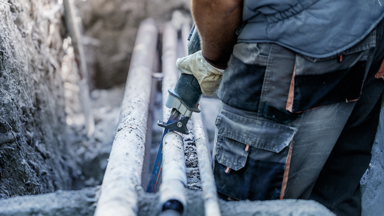 Seseorang yang mengenakan peralatan keselamatan memotong pipa dengan gergaji bolak-balik.