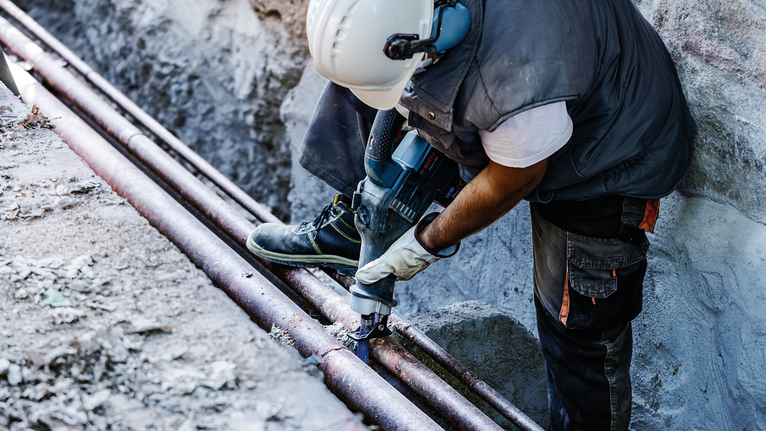 Seseorang yang mengenakan peralatan keselamatan menggunakan perkakas listrik untuk memotong pipa logam di luar ruangan.