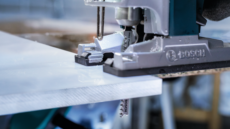 Mesin gergaji memotong lembaran plastik putih di sebuah bengkel.