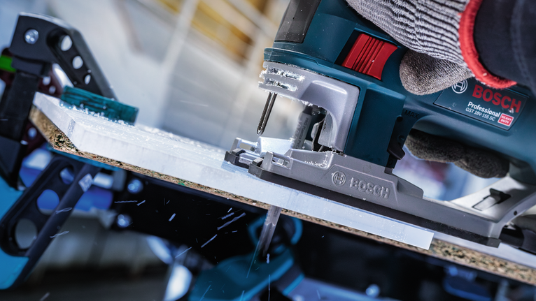 Seseorang yang mengenakan peralatan keselamatan mengoperasikan gergaji mesin untuk memotong selembar material.