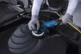 Seseorang yang mengenakan perlengkapan keselamatan memoles kap mobil dengan alat pemoles berwarna biru.