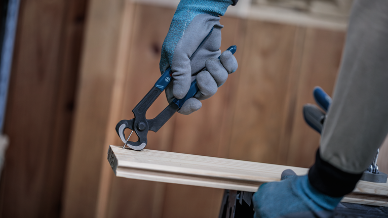 Seseorang yang mengenakan alat pelindung diri mencabut paku dari kayu menggunakan tang.