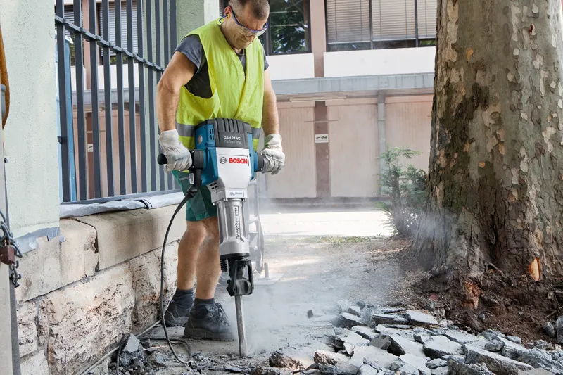 Seseorang yang mengenakan peralatan keselamatan merusak trotoar di luar ruangan dengan pemutus arus listrik berukuran besar.
