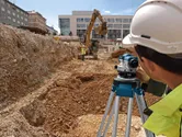 Orang yang mengenakan peralatan keselamatan menggunakan level surveyor di lokasi penggalian konstruksi.