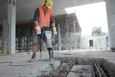 Seseorang yang mengenakan perlengkapan keselamatan menggunakan pemecah batu untuk menghancurkan beton di lokasi konstruksi.