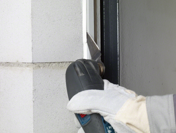 Seorang pekerja yang mengenakan peralatan keselamatan memotong lapisan penutup dari bingkai jendela dengan perkakas listrik.