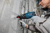 Seseorang yang mengenakan peralatan keselamatan mengebor dinding beton di sepanjang garis panduan biru.