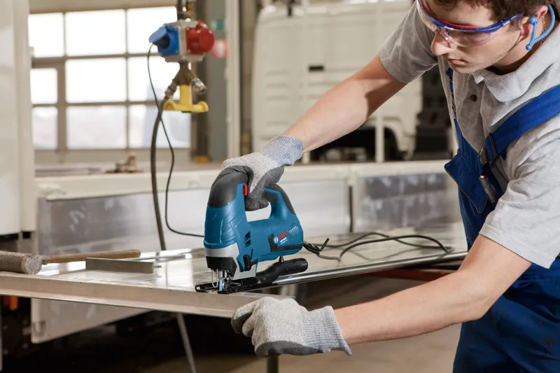 Seseorang yang mengenakan perlengkapan keselamatan menggunakan gergaji ukir untuk memotong lembaran logam.