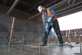 Seseorang yang mengenakan peralatan keselamatan mengebor beton dengan palu putar.